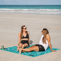 Two women sitting on a sand free teal beach blanket with ocean and sky in the background
