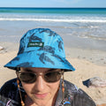 Lady wearing agreen/blue turtle bucket hat made by Amity Blue using recycled plastic bottles. The beach is in the background.