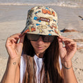 Lady wearing a beige retro kombi bucket hat made by Amity Blue using recycled plastic bottles. The beach is in the background.