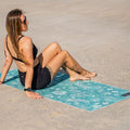 Woman sitting on a Amity Blue green and blue sand-free beach towel with a deep sea pattern on a sandy beach.