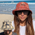 Woman wearing an orange patterned Amity blue bucket hat with sunglasses, standing on a beach.