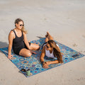 Two women sitting on a sand free blue Moroccan style beach blanket in the sand.