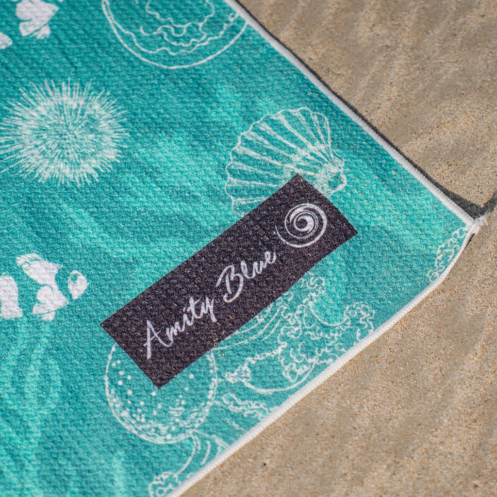 Teal sand free beach towel with sea creature patterns and 'Amity Blue' label on a concrete surface