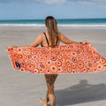 Woman on a beach holding an orange patterned sand free beach towel made from recycled plastic bottles with Amity Blue written on it