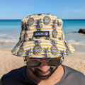 Man wearing a yellow and black pineapple bucket hat made by Amity Blue using recycled plastic bottles. The beach is in the background.