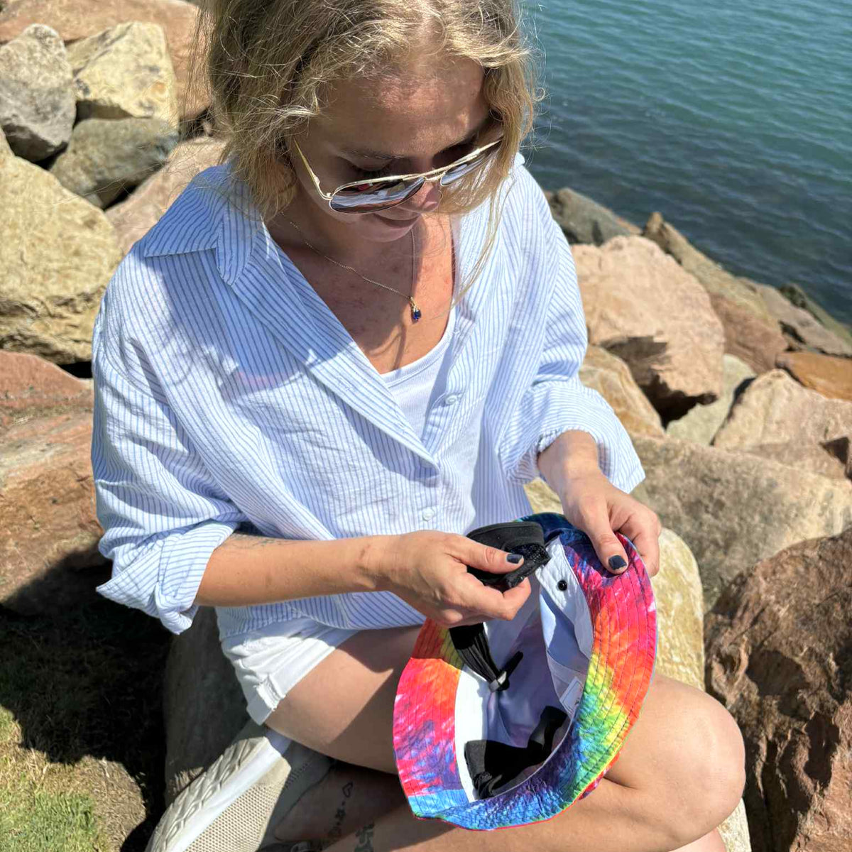 Person sitting on rocks by a body of water, holding a colorful tie dye hat, made from recycled plastic bottles by Amity Blue.