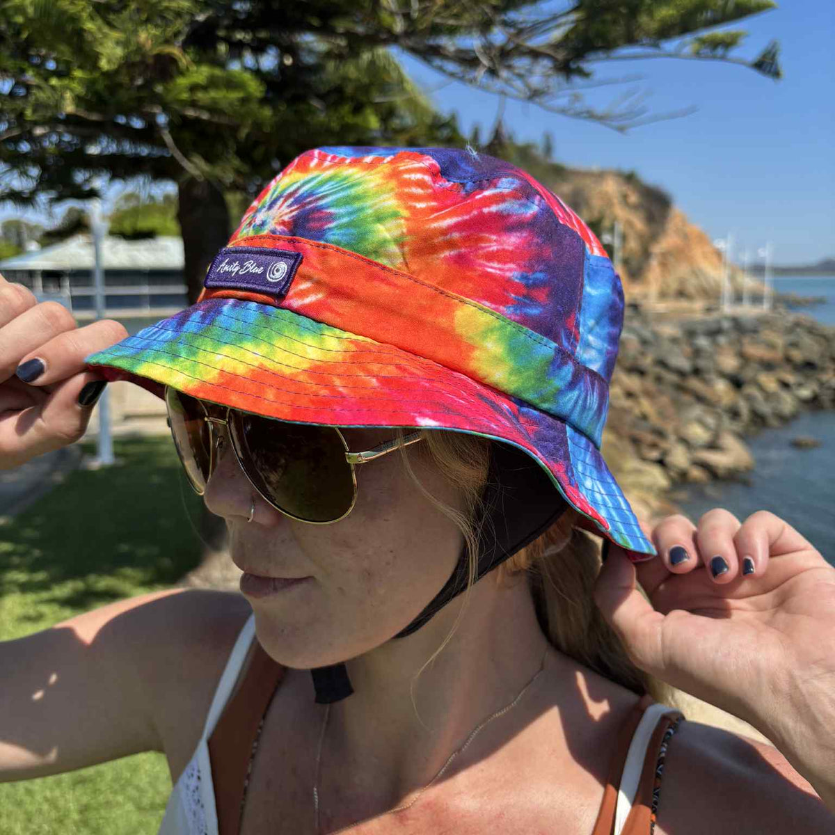 Person wearing a colorful tie-dye bucket hat with a scenic background. Amity Blue logo on hat. The hat is made from recycled plastic bottles