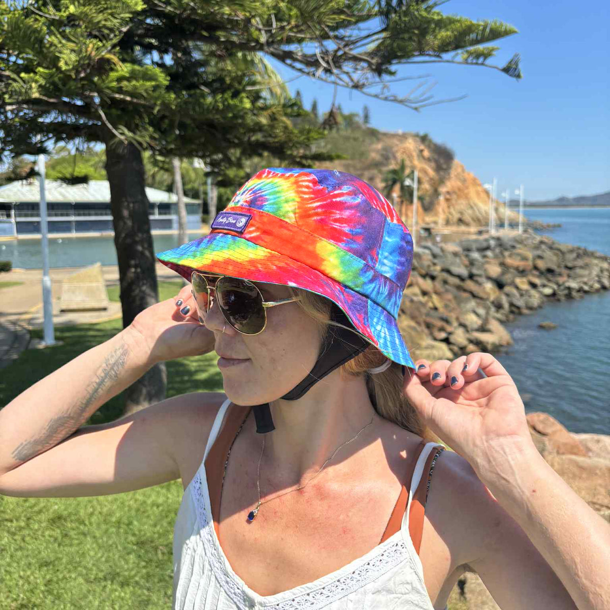 Person wearing a colorful tie-dye bucket hat by a waterfront with trees and rocks in the background. Hat is made by Amity Blue out of recycled plastic bottles.