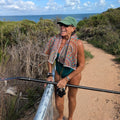 Woman standing on a trail by the ocean, holding a paddle board and wearing an Amity Blue India Go-To towel around her neck.